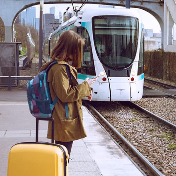 Frau steht mit gelbem Koffer am Bahnhof und Tram fährt ein