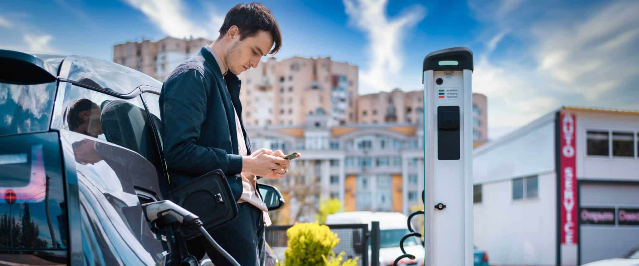 Ein junger Mann nutzt sein Handy während er sein E-Auto an der öffentlichen Ladestation lädt.