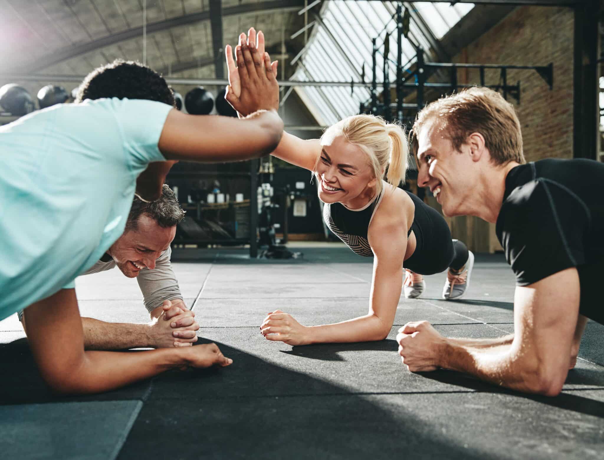 Drei Männer und eine Frau planken lächelnd gemeinsam im modernen Fitnessstudio, Frau und ein Mann geben sich High Five