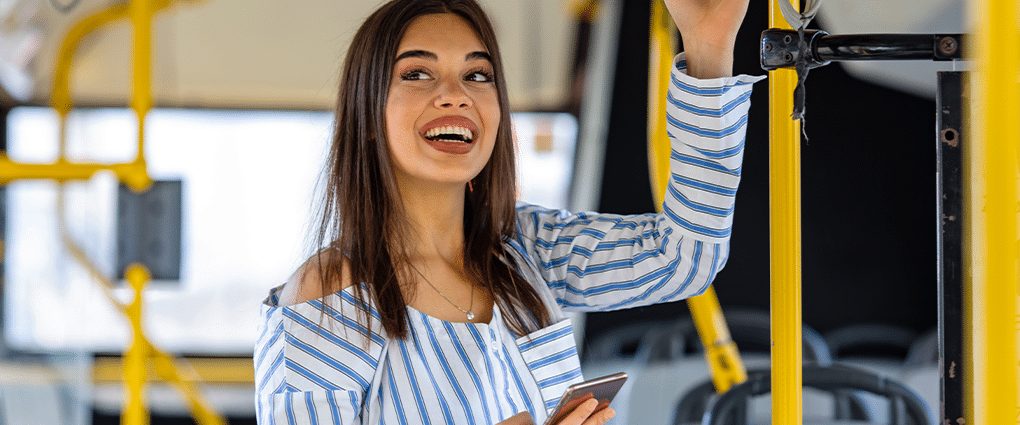 Junge, dunkelhaarige Frau schaut im Bus von ihrem Handy hoch.