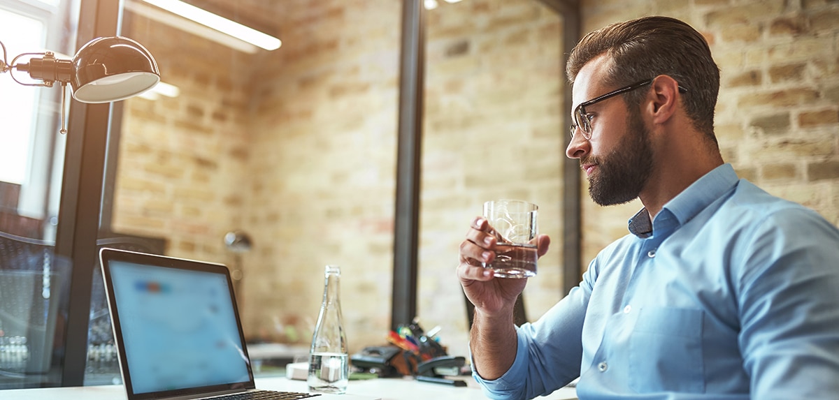 Mann mit Brille sitzt vor einem Laptop in einem modernen Büro und hält ein Glas Wasser.