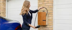Eine Frau mit blonden Haaren und Blazer hält ein Wallbox-Ladekabel neben ihrem Auto