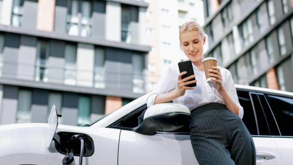Frau mit Handy und Coffee to go lehnt an weißem E-Auto