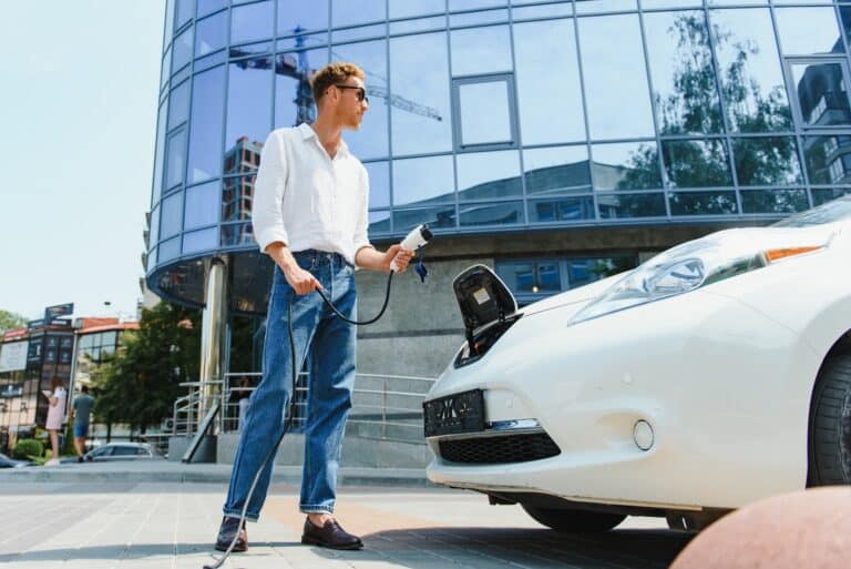 Ein Mann in weißem Hemd, Jeans und Sonnenbrille steht vor einem modernen Bürogebäude und lädt ein weißes Elektroauto, indem er ein Ladekabel in die Front des Fahrzeugs einsteckt.