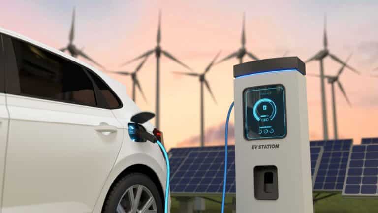 Weißes Auto lädt an Wallbox, dahinter, Windpark und PVC-Anlage